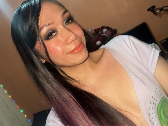 TsJade has long black and purple hair, big green eyes, and glossy pink lips. She's wearing a white top with a green design, showing her small tits.