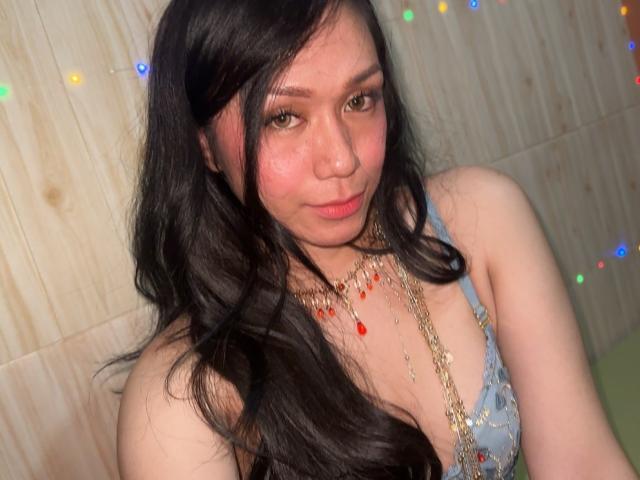 TsJade with long black hair, wearing a blue lace bra, gold chain necklace with red beads, and a slight smile, looking at the camera, showing her tits.