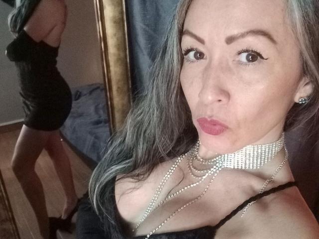 NicoletteShea with long gray hair, wearing a black dress, pearl necklace, and choker, pouting her lips, standing in front of a mirror showing her ass.