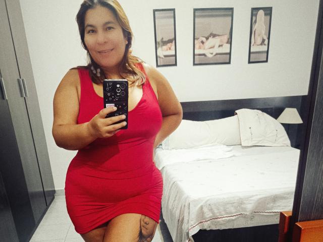 LouiseSexBigBoobs, curvy with big tits, wearing a tight red dress, standing in a bedroom, taking a mirror selfie, showing a tattoo on her thigh, smiling.