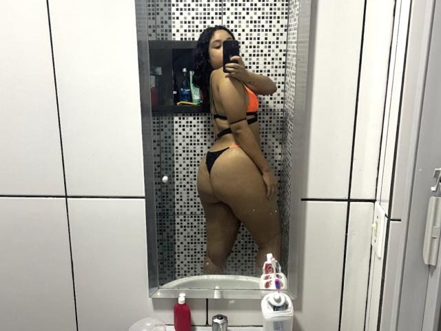 RobertinhaCandy with black thong and orange bra, showing her curvy ass and thick thighs, taking a mirror selfie in a bathroom, her dark hair falling to the side.