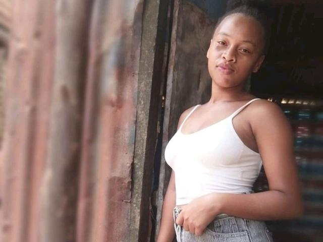 ISmileLove with medium brown skin, small tits in a white tank top, and high-waisted jeans, standing by a rusty wooden door, looking slightly to the side.