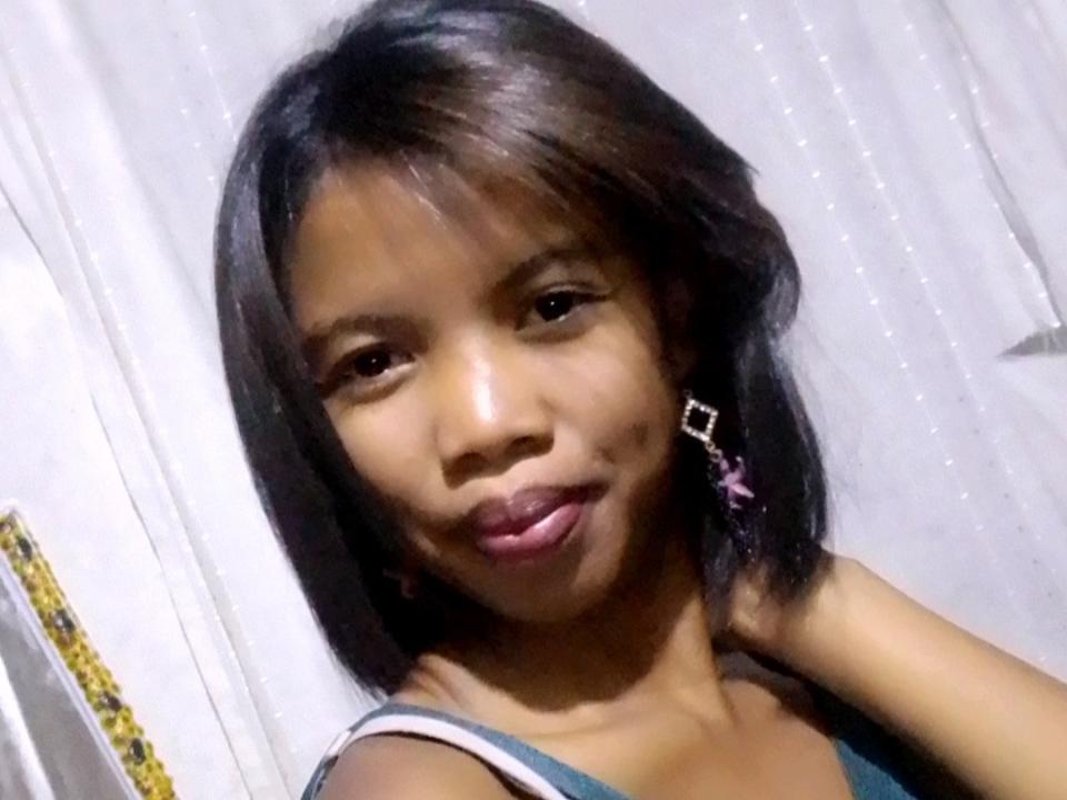 MendrikaSweet with medium brown skin, black shoulder-length hair, small purple earrings, and slightly puffed lips, wearing a blue top. Her tits are small, and her face is cute.