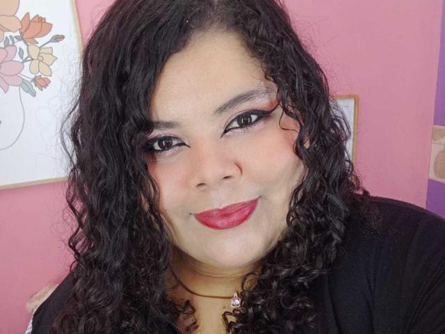 SarahBowie has curly black hair, red lipstick, and eyeliner, smiling at the camera. She's wearing a black top and a necklace with a small heart. Pink wall in background.