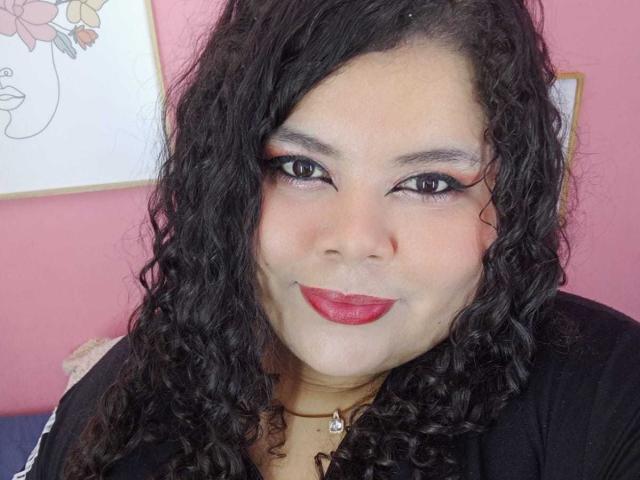 SarahBowie with curly black hair, red lipstick, and black eyeliner, smiling at the camera, wearing a black top with a gold necklace, pink walls in the background.