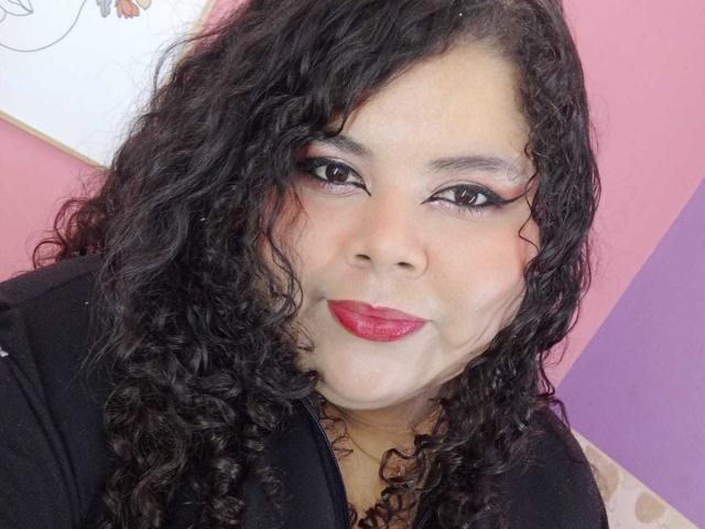SarahBowie has curly black hair, red lipstick, and black eyeliner. She's wearing a black top, looking directly at the camera with a slight smile. Her skin is light brown.