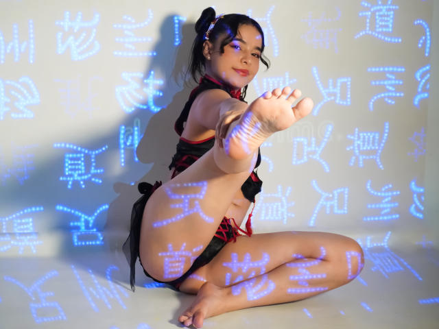 LissyCarter with black and red lingerie, kneeling, showing her bare ass and foot, dark hair in puffy ponytail, blue light Chinese characters on white wall, smiling.