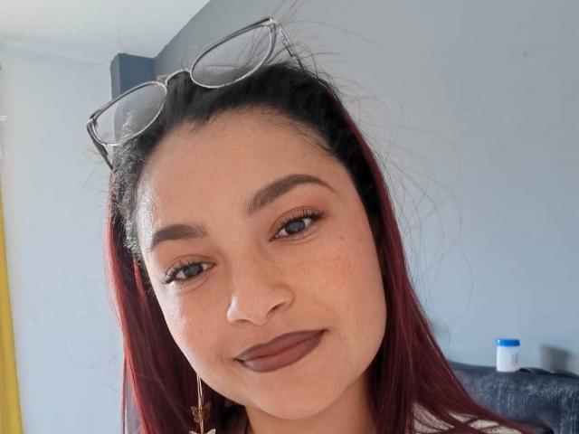 Ameliebx a les cheveux d'un rouge foncé, porte des lunettes sur la tête, du fard à paupières marron et du rouge à lèvres foncé. Elle sourit et a des taches de rousseur sur le visage. Sa peau, d'un brun moyen, est visible..