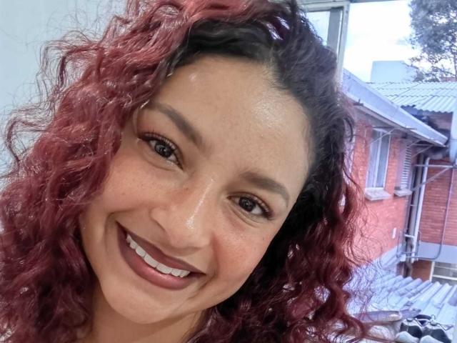 Ameliebx, avec ses cheveux roux-brun bouclés, sourit, montrant des dents blanches, a des yeux marron et une peau brun clair, a un visage mignon et porte un rouge à lèvres brun foncé..