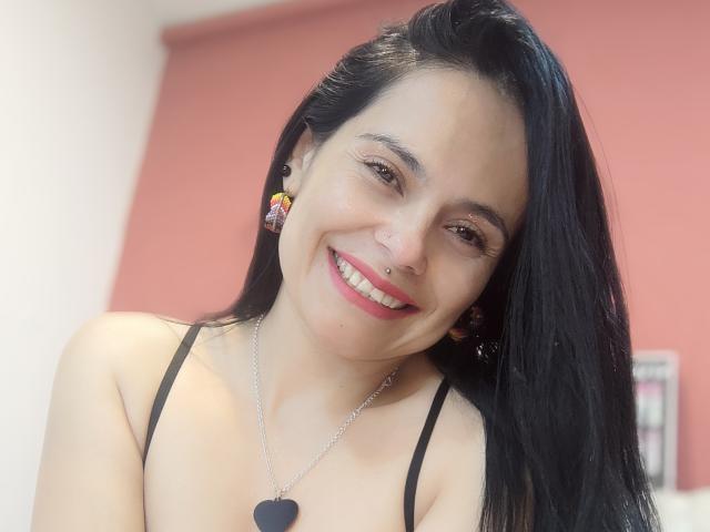 CharlotteCrofort with long black hair, smiling, wearing a black strap top, nose ring, and heart necklace, showing her tits, pink lipstick, and colorful earrings.