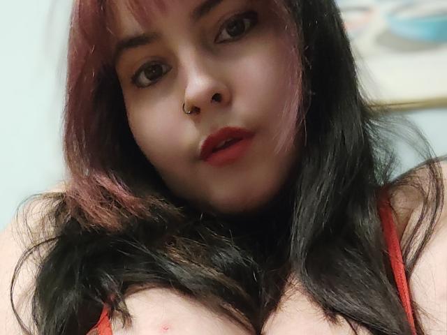 SelenaBoni has dark, wavy hair with red highlights, red lipstick, and a nose ring. Her red tank top reveals her large, bare tits with a nipple visible.