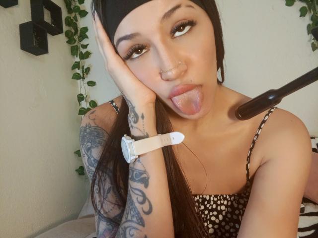 AnnaKhalo with black headscarf, tongue out, hand on cheek, tattooed arm, white wristwatch, black and white striped top, and nose ring, wearing a microphone on a stand.