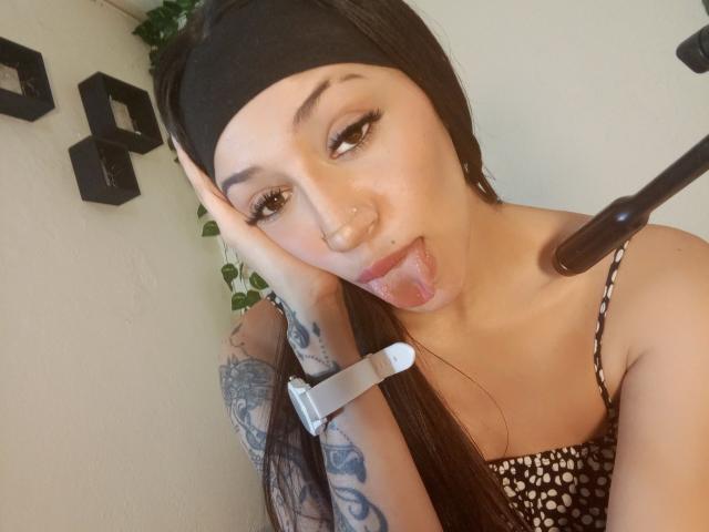 AnnaKhalo with long black hair, black headband, and tattooed arm, tongue out, holding phone to camera, wearing a polka-dot top, and a white wristband.