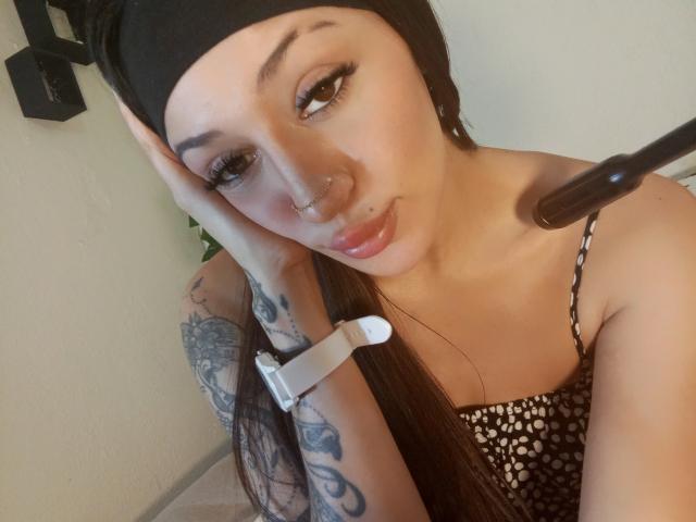 AnnaKhalo with black headscarf, long brown hair, nose ring, and lip ring, looking at camera, wearing black and white polka dot top, tattoo on right arm, wristband on left.