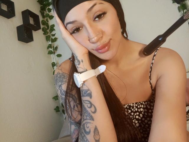 AnnaKhalo with long brown hair in a black beanie, tattooed arm, nose ring, and white smartwatch, wearing a black and white polka dot top, smiling at the camera.