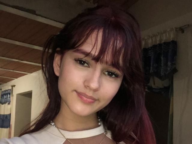 MelodyMansson has red hair, green eyes, and a cute smile with subtle makeup. She wears a white shirt and a necklace, showing a bit of her tits.