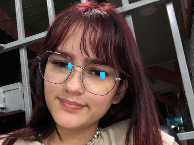 MelodyMansson with red-brown hair, glasses reflecting blue light, smiling, wearing a pearl necklace, light skin, and a beige top. Her tits are not visible.