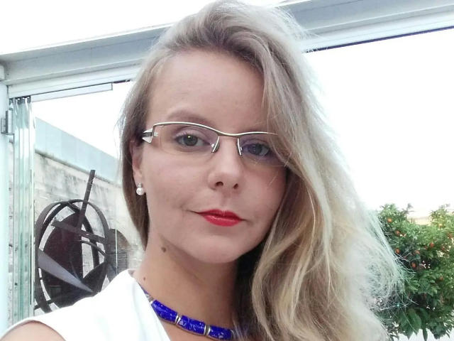 NylonPussy20-hot has blonde hair, red lipstick, glasses, white shirt, blue necklace, pearl earrings, and a small mole on her chin. Her ass is not visible.