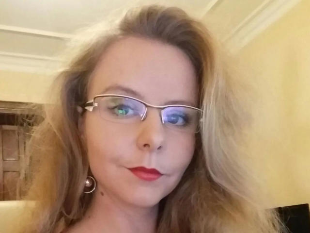 NylonPussy20-hot has blonde hair, red lipstick, and wears glasses with metal earbuds. Her blue eyes are slightly crossed, giving a quirky look. She's smiling subtly.