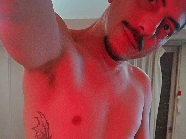 Shirtless XSantyX with a red glow, mustache, and goatee, showing a tattoo on his left side, taking a selfie. His arm is raised, and he's smiling slightly.