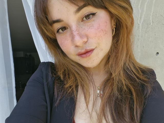 AlennaVolt has freckled, fair skin with light brown, wavy hair. She wears a black top, showing small tits, with a necklace and gold earrings, smiling softly.