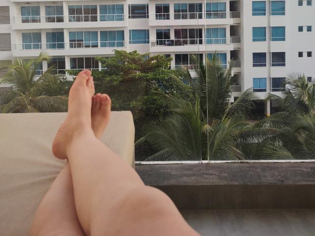 AlennaVolt's legs are crossed at the ankles, showing her bare feet with pink toenails, resting on a beige couch. Tall building with blue windows in the background.