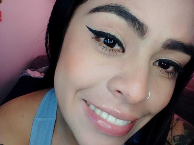 StellaBeckker's face with dark hair, light skin, thick eyebrows, blue eyes, black eyeliner, nose ring, glossy pink lips, smiling, wearing a blue top, visible shoulder.