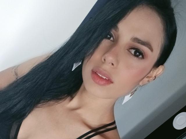 AnaisHilton with long black hair, brown eyes, and pink lips, wearing black straps and white triangle earrings, showing her smooth, cute face and subtle tattoo on her arm.
