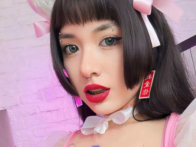 SabriPrettty has a cute face with dark brown bob, pink bows, red lipstick, nose piercing, and white frilled choker. She's wearing pink and white, with red earrings.
