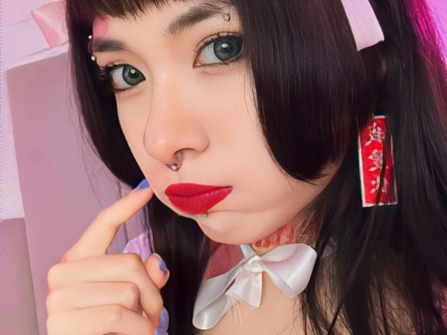 SabriPrettty has pale skin, green eyes, black hair with pink highlights, nose piercings, red lipstick, blue nails, white bow, and red earrings, touching her lips with a finger.