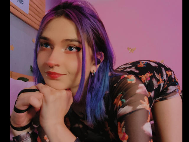 MaiaThunder met paars-blauw haar, een zwarte bloemenjurk en rode lippenstift, leunt op haar hand, glimlachend, en laat haar neusring en oorbellen zien, met haar billen iets omhoog..