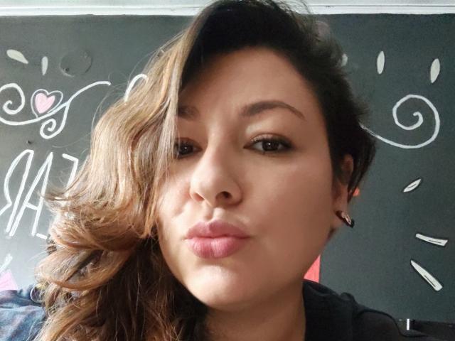 SakotoDika hat braunes, welliges Haar, hellbraune Haut und zieht die Lippen zu einem Schmollmund, wobei pinkfarbene Lippenstift sichtbar ist. Sie trägt kleine Ohrringe und ein schwarzes Hemd. Ihre Augen sind leicht zusammengekniffen..