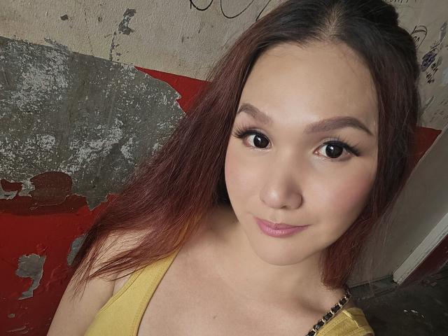 LovelyPinay69 with long red-brown hair, pink lipstick, and thick eyelashes, wearing a yellow top, showing her cute face and small tits. Her skin is light and she's smiling.