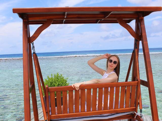 SafiraFuracao, com seios pequenos, vestindo um vestido branco e óculos de sol, sentada em um gazebo de madeira, com a mão na cabeça, olhando para a câmera, com um oceano azul claro e céu ao fundo..