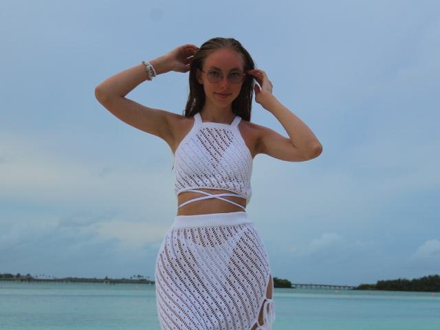 SafiraFuracao, con lunghi capelli castani, indossa un top e una gonna bianchi in rete, è in piedi vicino all'oceano, con le mani tra i capelli, sullo sfondo c'è un cielo azzurro e acqua turchese..