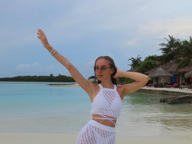 SafiraFuracao com os braços levantados, vestindo uma blusa cropped branca e transparente de malha e uma saia combinando, em pé em uma praia com água cristalina e palmeiras ao fundo..