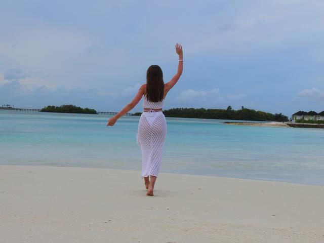 SafiraFuracao draagt een witte jurk van gaas, staat met haar rug naar de camera, haar armen omhoog, en loopt zonder schoenen op een strand met turquoise water en een bewolkte lucht. Haar achterkant is zichtbaar..