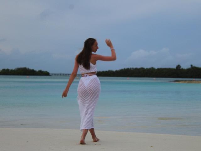 SafiraFuracao, avec de longs cheveux bruns, un haut blanc sans manches et une jupe blanche transparente, agitant la main sur une plage, avec de l'eau bleu-vert et un ciel nuageux en arrière-plan..