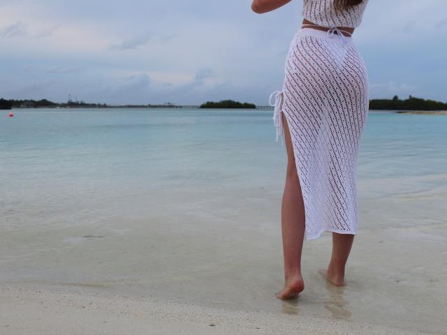 SafiraFuracao trägt einen weißen, durchsichtigen, gehäkelten Rock mit einem seitlichen Schlitz und steht barfuß im Wasser am Strand, wobei ihr Hintern zu sehen ist..