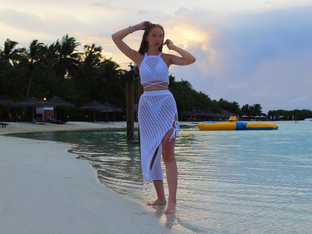 SafiraFuracao in piedi su una spiaggia, indossa un top e una gonna bianchi in rete, con le mani tra i capelli, acqua blu, un oggetto gonfiabile giallo, palme, tramonto e sabbia bianca..