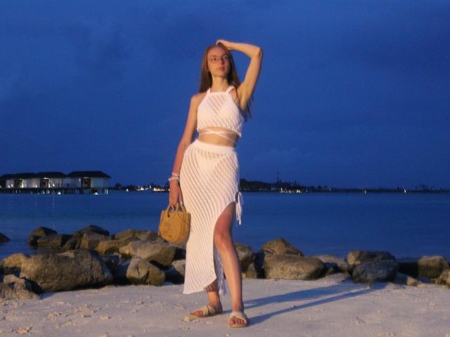 SafiraFuracao se tient sur une plage au crépuscule, vêtue d'un ensemble blanc et transparent en deux pièces avec une fente haute, tenant un sac en osier, sa main sur sa tête..