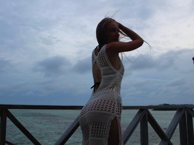 SafiraFuracao portant une robe blanche en crochet avec une fente haute, dévoilant ses fesses, debout sur un balcon, une main dans ses cheveux, avec un ciel nuageux en arrière-plan..