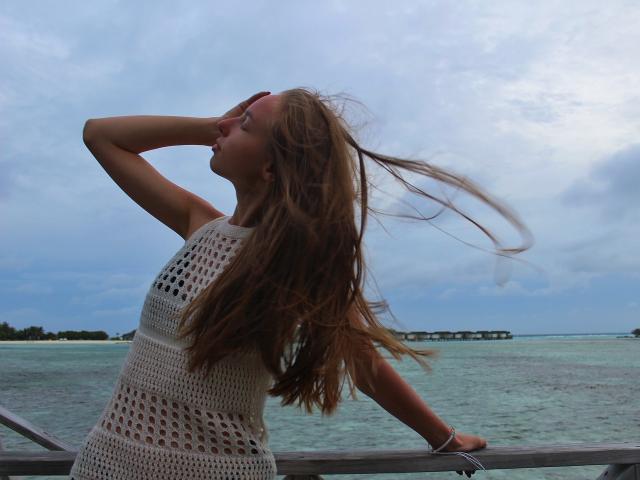 SafiraFuracao con lunghi capelli castani, un vestito bianco all'uncinetto, una mano davanti agli occhi, appoggiata a una ringhiera di legno, con il vento che le scompiglia i capelli. In secondo piano, si vedono l'oceano e un cielo nuvoloso. Le sue natiche sono leggermente visibili..