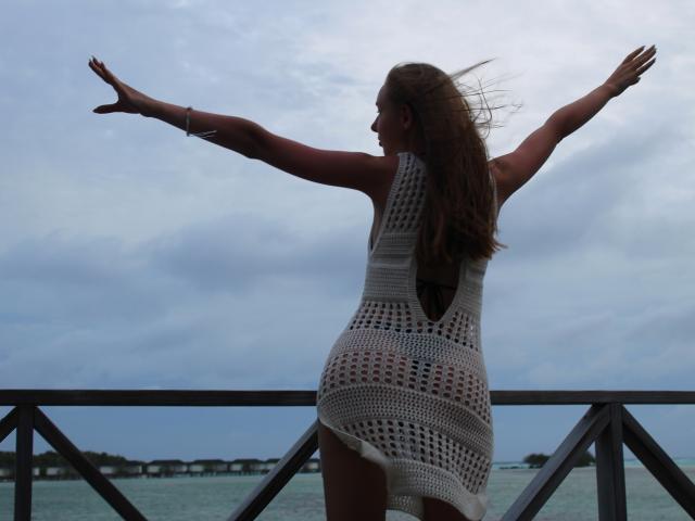 SafiraFuracao está de pie con los brazos extendidos, de espaldas a la cámara, vistiendo un vestido blanco de crochet, mostrando su trasero, sobre una barandilla de metal, cerca del agua, con el pelo castaño ondeando por el viento..