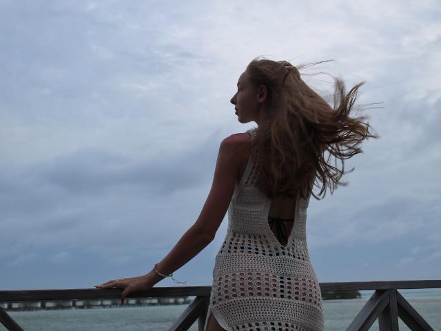 SafiraFuracao, avec de longs cheveux bruns, portant une robe blanche en crochet, debout dos à la caméra, les mains sur une rambarde, regardant sur le côté, ciel nuageux..