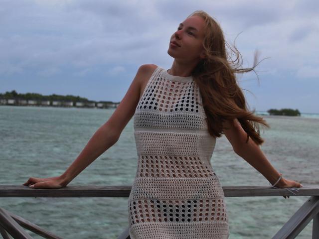SafiraFuracao, con lunghi capelli castani, indossa un abito bianco all'uncinetto, appoggiata a una ringhiera, con l'oceano sullo sfondo, guarda verso l'alto con un leggero sorriso, con le mani appoggiate alla ringhiera..