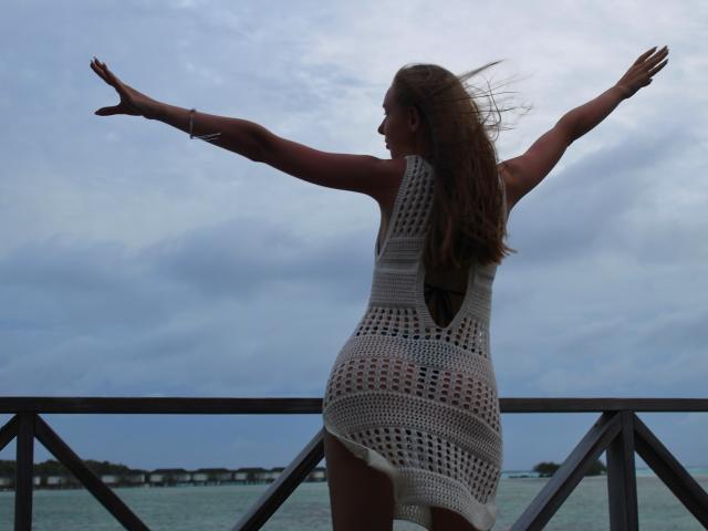 SafiraFuracao está de pie con los brazos extendidos, vistiendo un vestido blanco de crochet, mostrando su trasero, frente a un cielo y mar nublados, con su cabello castaño ondeando..