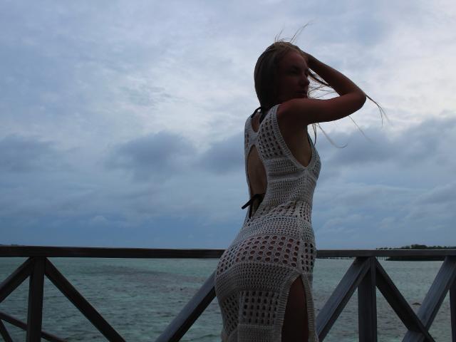 SafiraFuracao vestindo um vestido branco de crochê com uma fenda lateral, mostrando suas nádegas e costas, em pé em uma grade de metal à beira-mar, com a mão no cabelo..