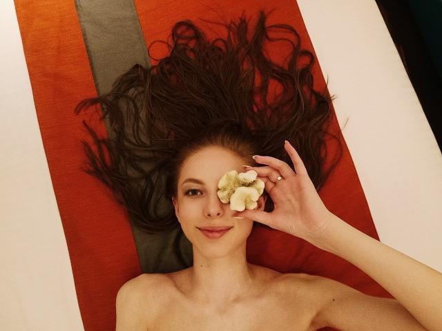 SafiraFuracao allongée en bikini sur une couverture rouge et grise, cachant un œil avec une fleur blanche et verte, de longs cheveux bruns éparpillés, souriant doucement..