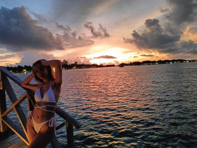 SafiraFuracao en maillot de bain blanc, debout sur une jetée en bois au coucher du soleil, les mains dans les cheveux, l'eau reflétant un ciel orange et violet, une image calme et sensuelle..